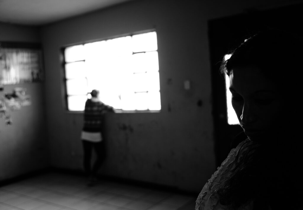 black-and-white photo; scene depicts aftermath of domestic violence. One silhouette is in the foreground; another is in the background, showing a woman looking out a large window.